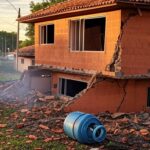 Residência em Ceilândia danificada por explosão de botijão de gás, com destroços e fumaça, em estilo fotojornalístico.