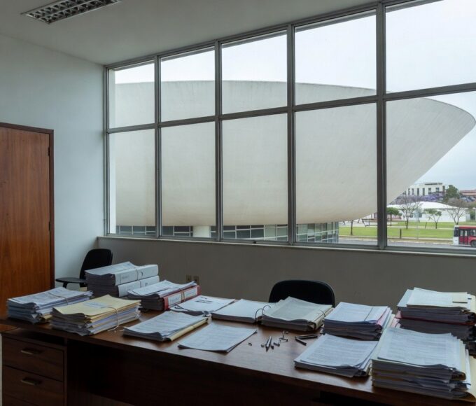 Edifício moderno em Brasília com pilhas de documentos burocráticos, ilustrando exclusão de associações de consumidores pela Caesb e Adasa.