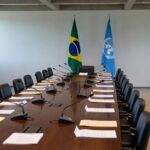 Sala de conferência em Brasília com bandeiras do Brasil e ONU, representando críticas a guerras, fome e descrédito da ONU.