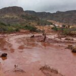 Paisagem rural na Zona da Mata Mineira após tragédia, com casas danificadas e rio transbordado, representando vítimas de desastre natural.