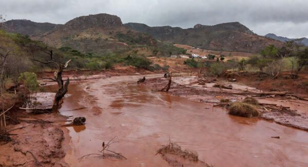 Paisagem rural na Zona da Mata Mineira após tragédia, com casas danificadas e rio transbordado, representando vítimas de desastre natural.