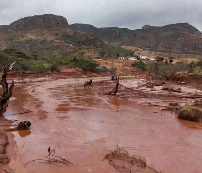 Paisagem rural na Zona da Mata Mineira após tragédia, com casas danificadas e rio transbordado, representando vítimas de desastre natural.