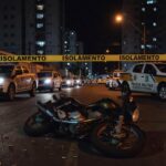 Cena de crime em rua de Taguatinga, DF, com fita de isolamento, motocicleta abandonada e marcas de tiros no asfalto.