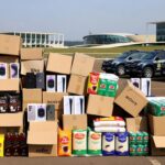 Mercadorias irregulares apreendidas em operação no DF, com caixas empilhadas e viaturas policiais ao fundo.