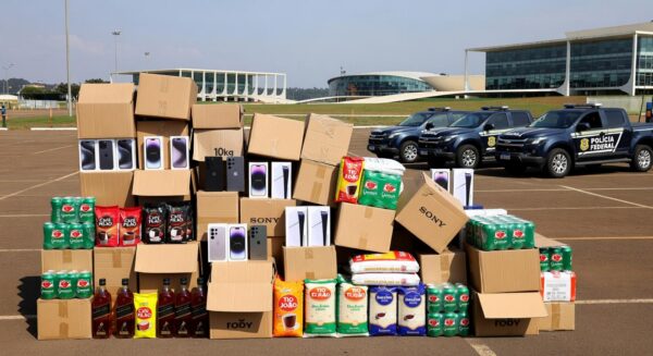 Mercadorias irregulares apreendidas em operação no DF, com caixas empilhadas e viaturas policiais ao fundo.