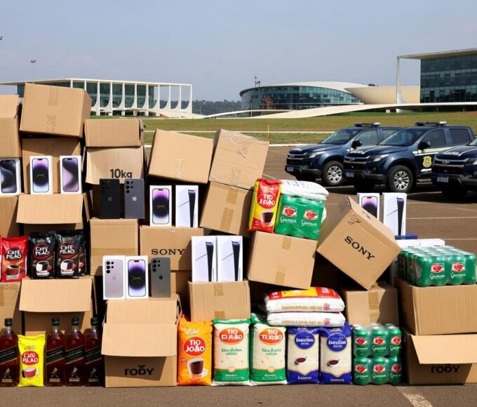 Mercadorias irregulares apreendidas em operação no DF, com caixas empilhadas e viaturas policiais ao fundo.